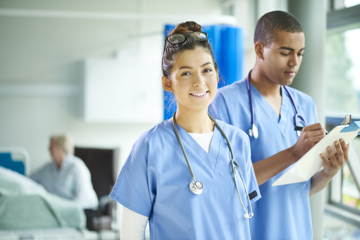 a young male and female nurse stand proudly on the ward and look to camera . In the background a patient is being attended by a nurse and their relative .