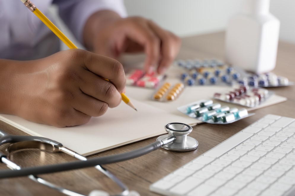 A health professional at a desk writing a note, with different packets of tablets and medication on the desk.